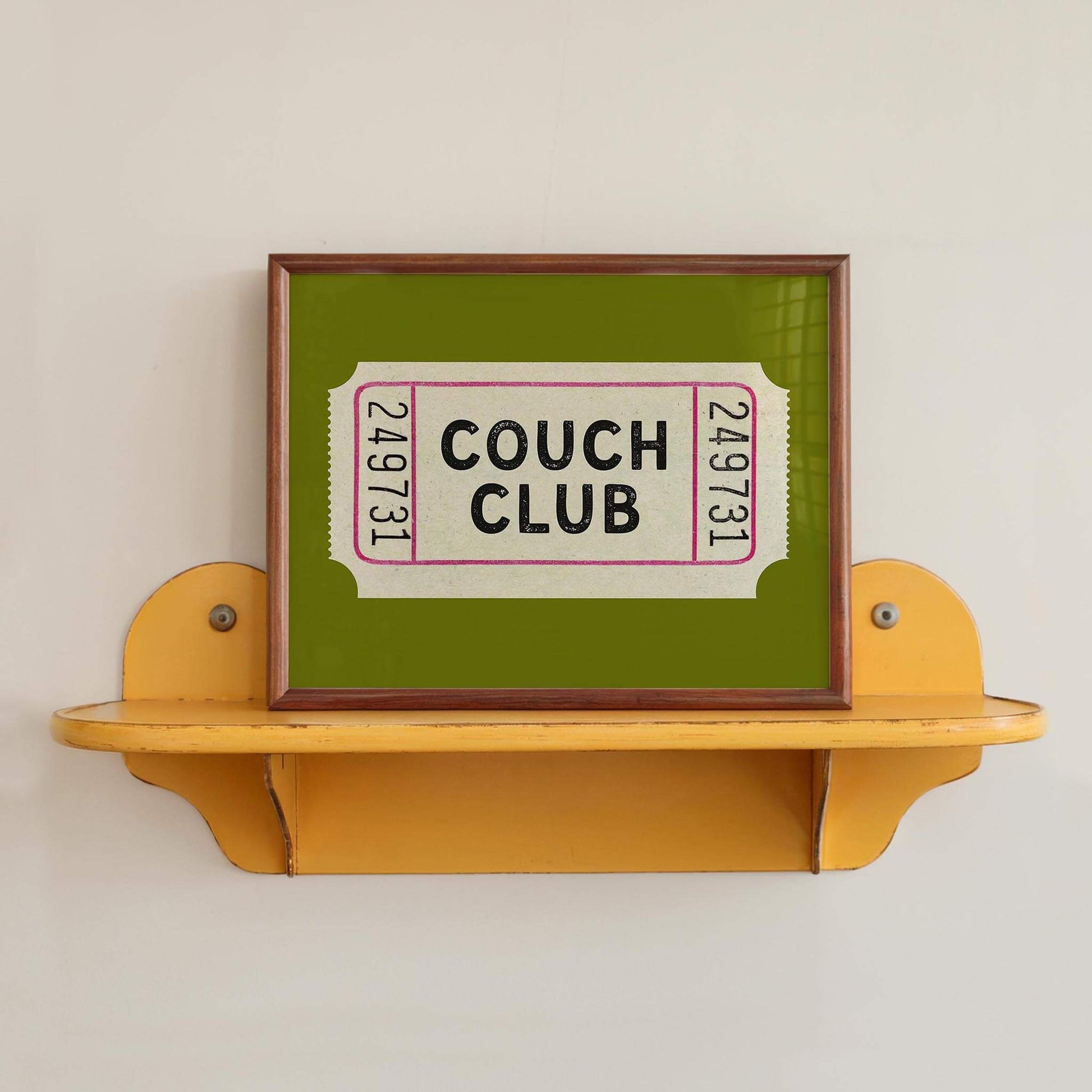 A framed image of a green ticket with the text "Couch Club" displayed on it, placed on a yellow shelf against a white wall.