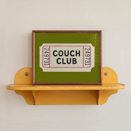 A framed image of a green ticket with the text "Couch Club" displayed on it, placed on a yellow shelf against a white wall.