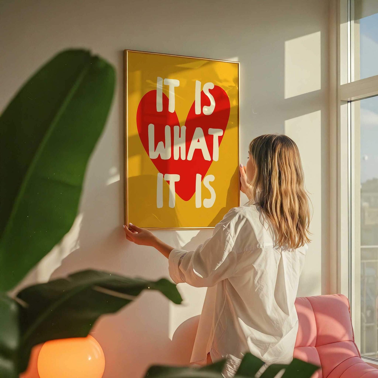 A woman hangs a yellow poster with the text "IT IS WHAT IT IS" on a wall in a room with a pink couch and a plant.