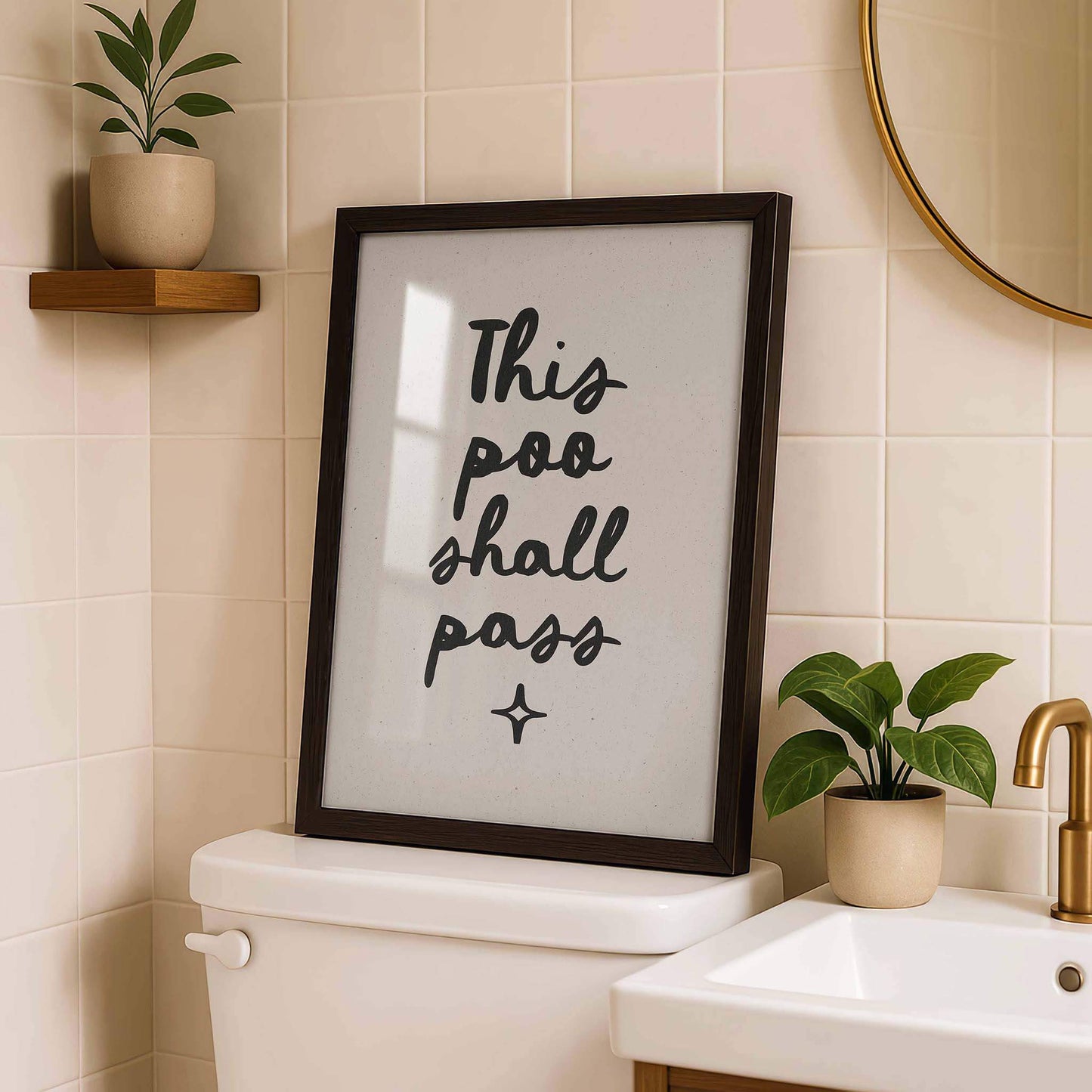 a framed sign with the text "This poo shall pass" in a bathroom setting, accompanied by a potted plant and a sink.