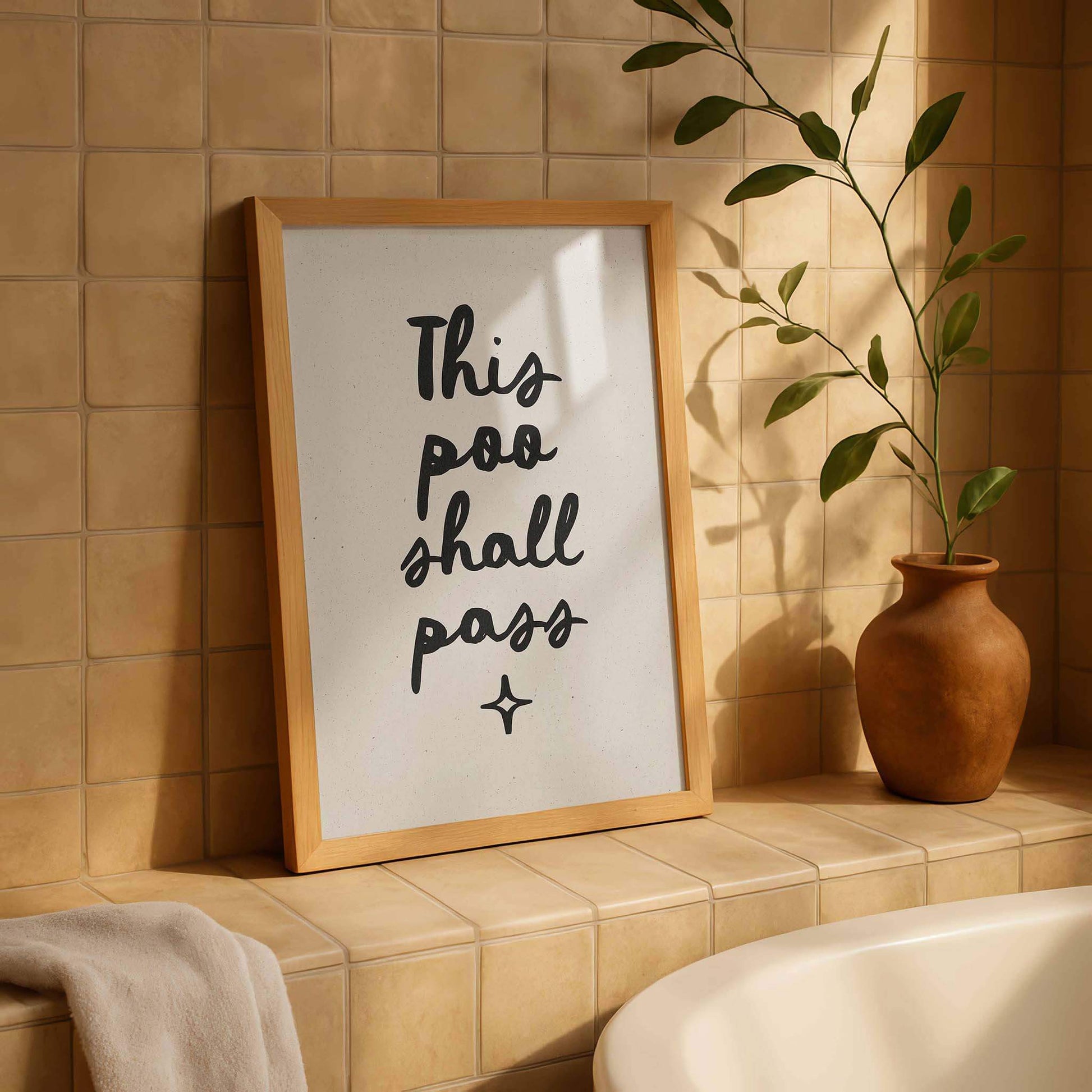 a framed sign with the text "This poo shall pass" in a wooden frame, placed on a tiled wall next to a potted plant.