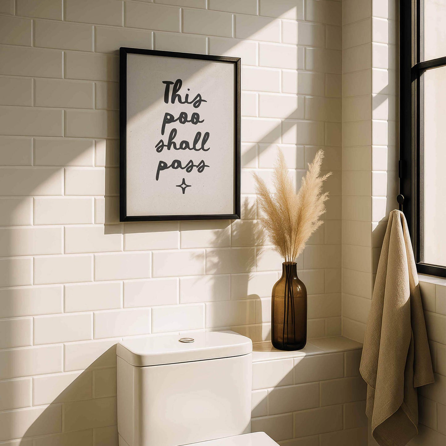 a bathroom with a framed sign that reads "This poo shall pass" above a toilet. The bathroom has white tiles and a window, and there is a vase with dried flowers on the windowsill.