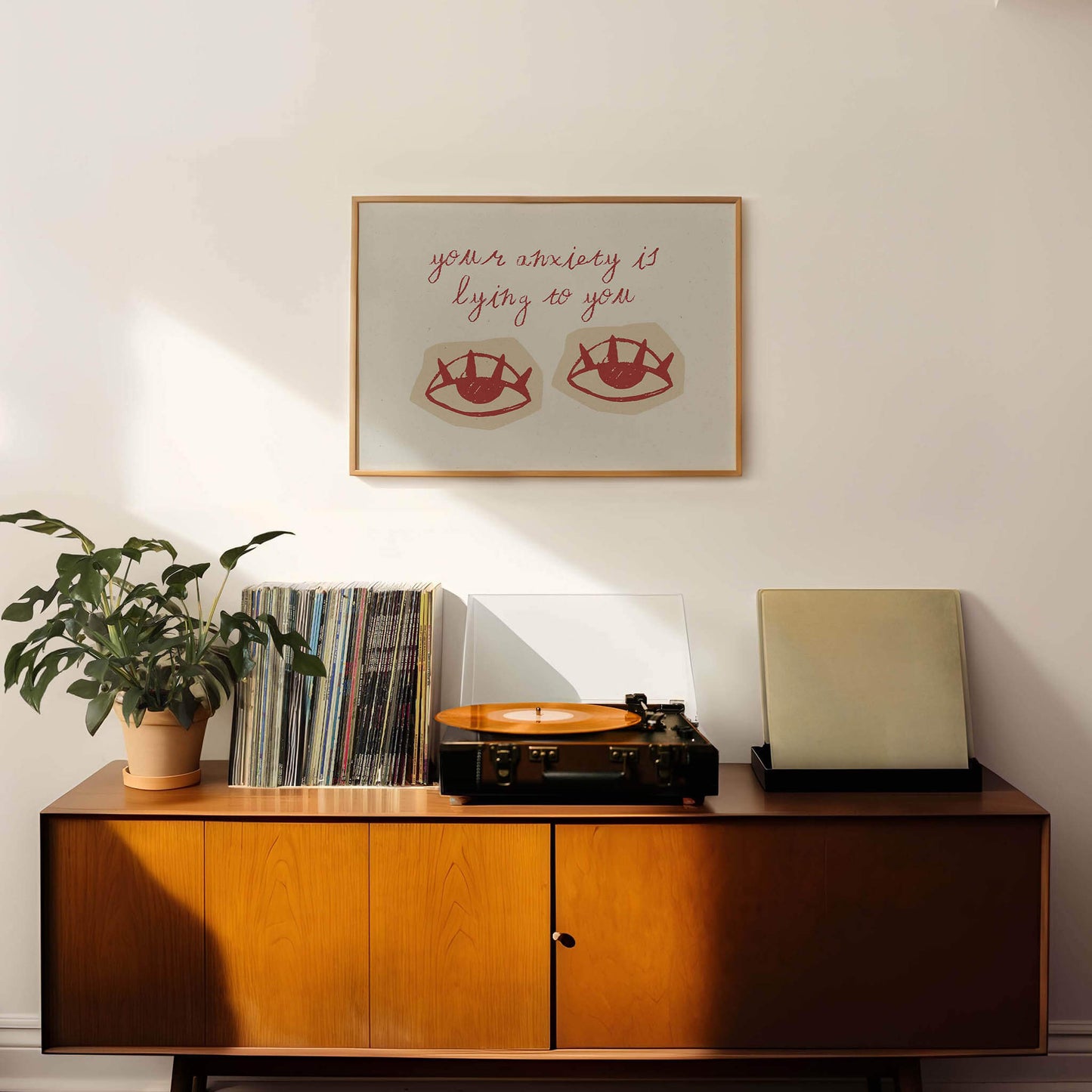 a wooden cabinet with a record player and a stack of vinyl records on top. Above the cabinet, there is a framed quote that reads "your anxiety is lying to you".