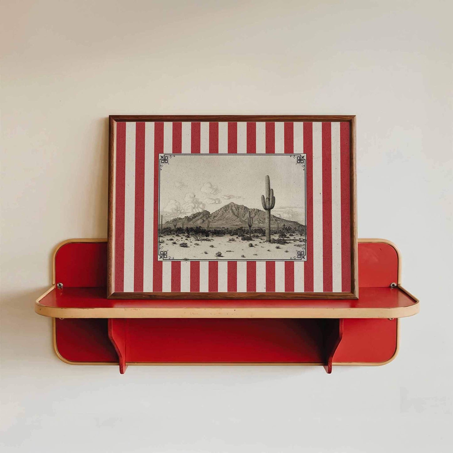 a framed picture of a desert landscape with a cactus in the center, mounted on a red shelf against a white wall.