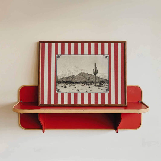 a framed picture of a desert landscape with a cactus in the center, mounted on a red shelf against a white wall.