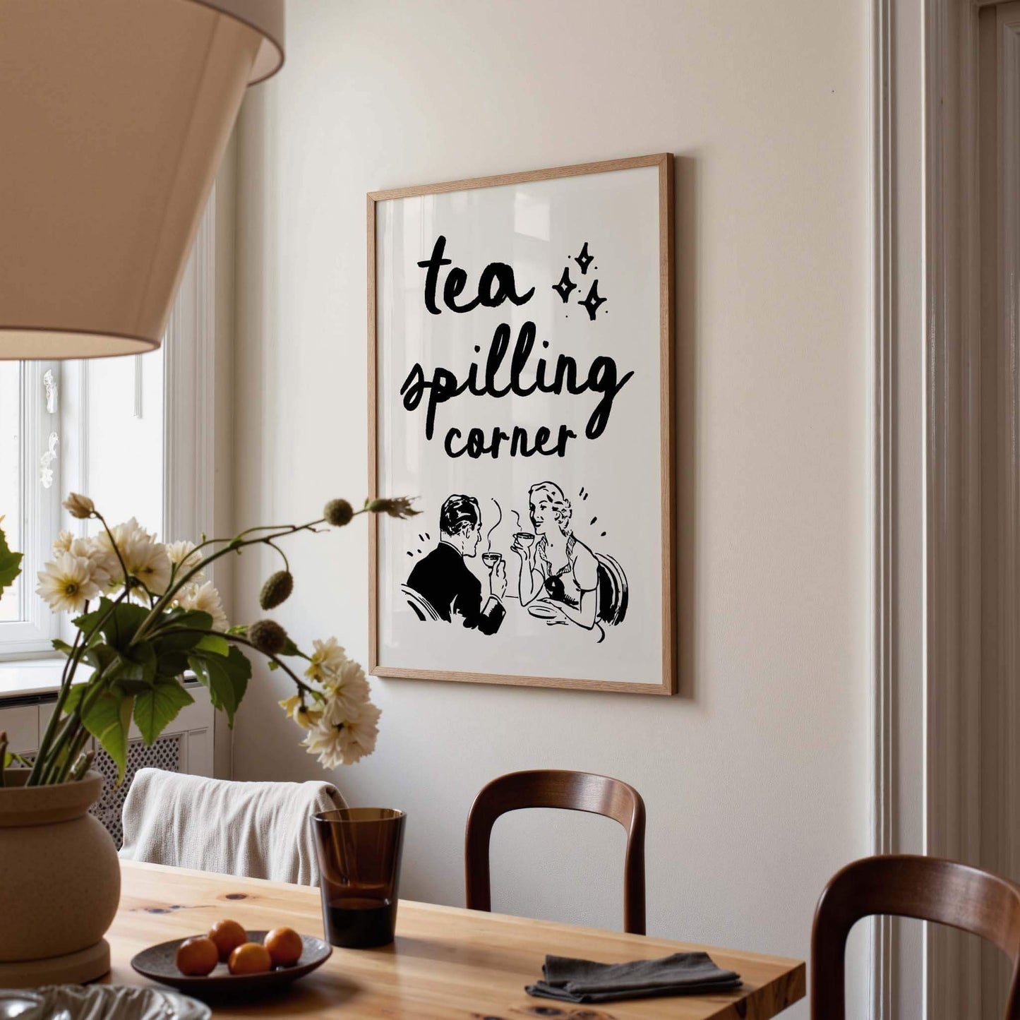 a framed poster with the text "tea spilling corner" hanging on a wall above a wooden dining table with chairs.