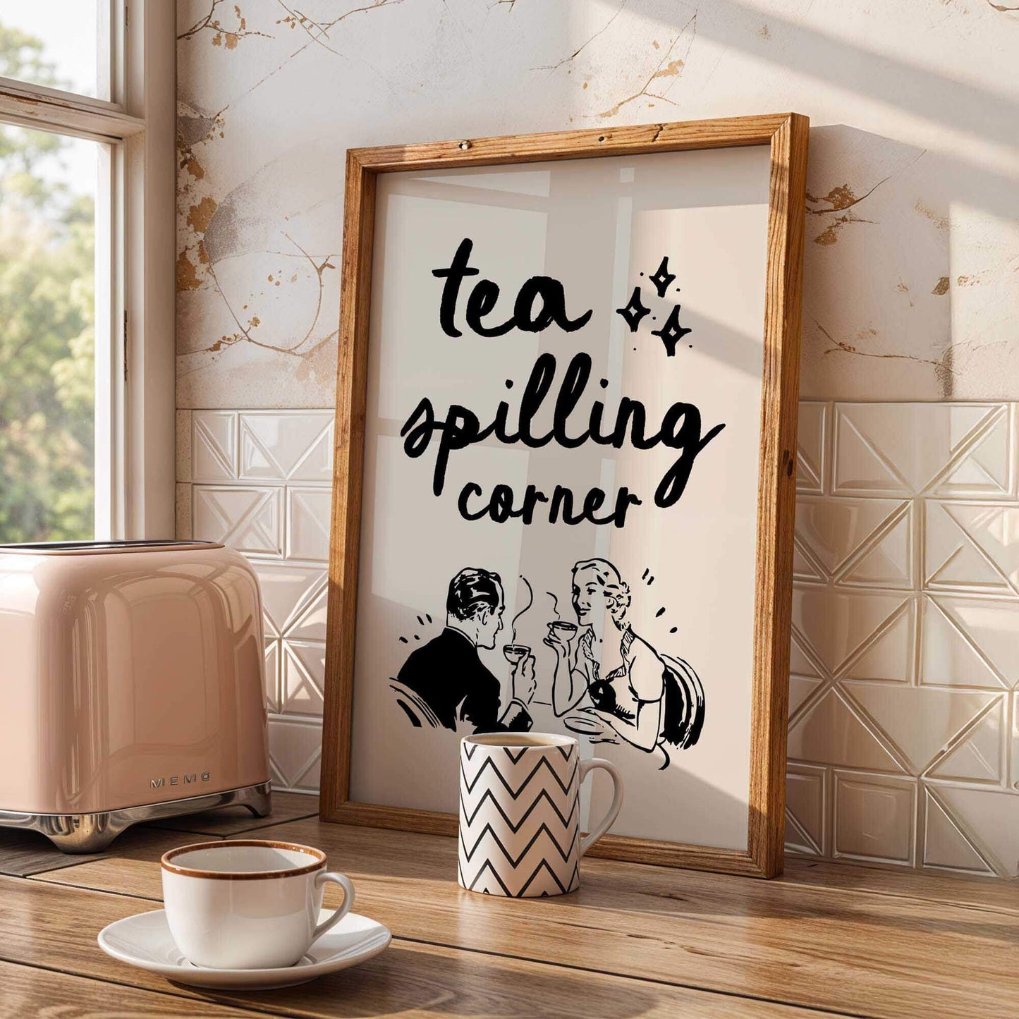 a framed poster with a vintage-style illustration of a couple sitting at a table, accompanied by the text "tea spilling corner" in a playful, retro font. The poster is displayed on a kitchen counter, with a toaster and a coffee cup visible in the foreground.
