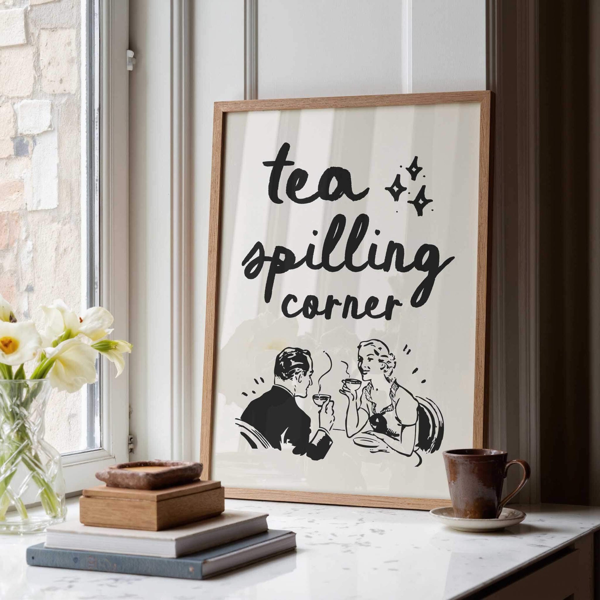 a framed poster with the text "tea spilling corner" and an illustration of a man and a woman sitting at a table, enjoying tea together. The poster is displayed on a white wall, and there is a vase of white flowers and a stack of books on a table nearby.