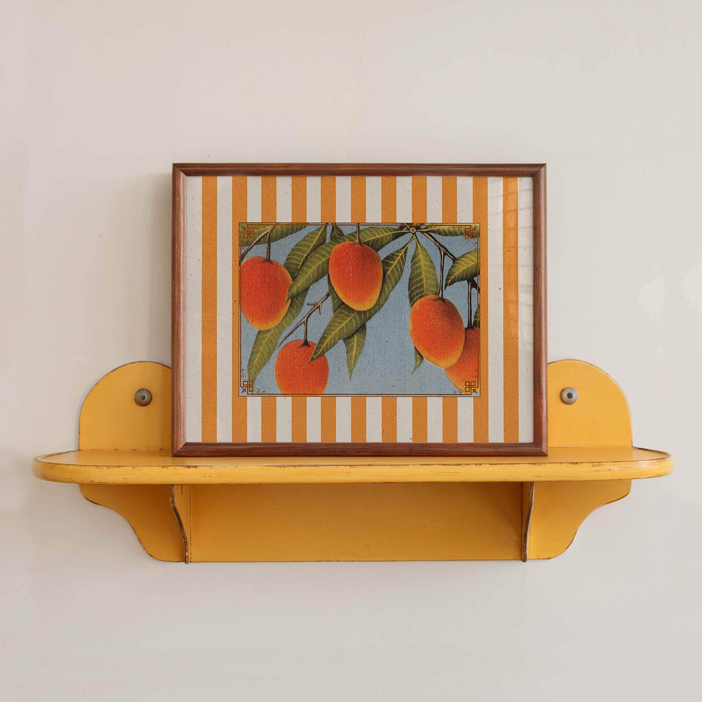 A framed painting of oranges is displayed on a yellow shelf against a white wall.