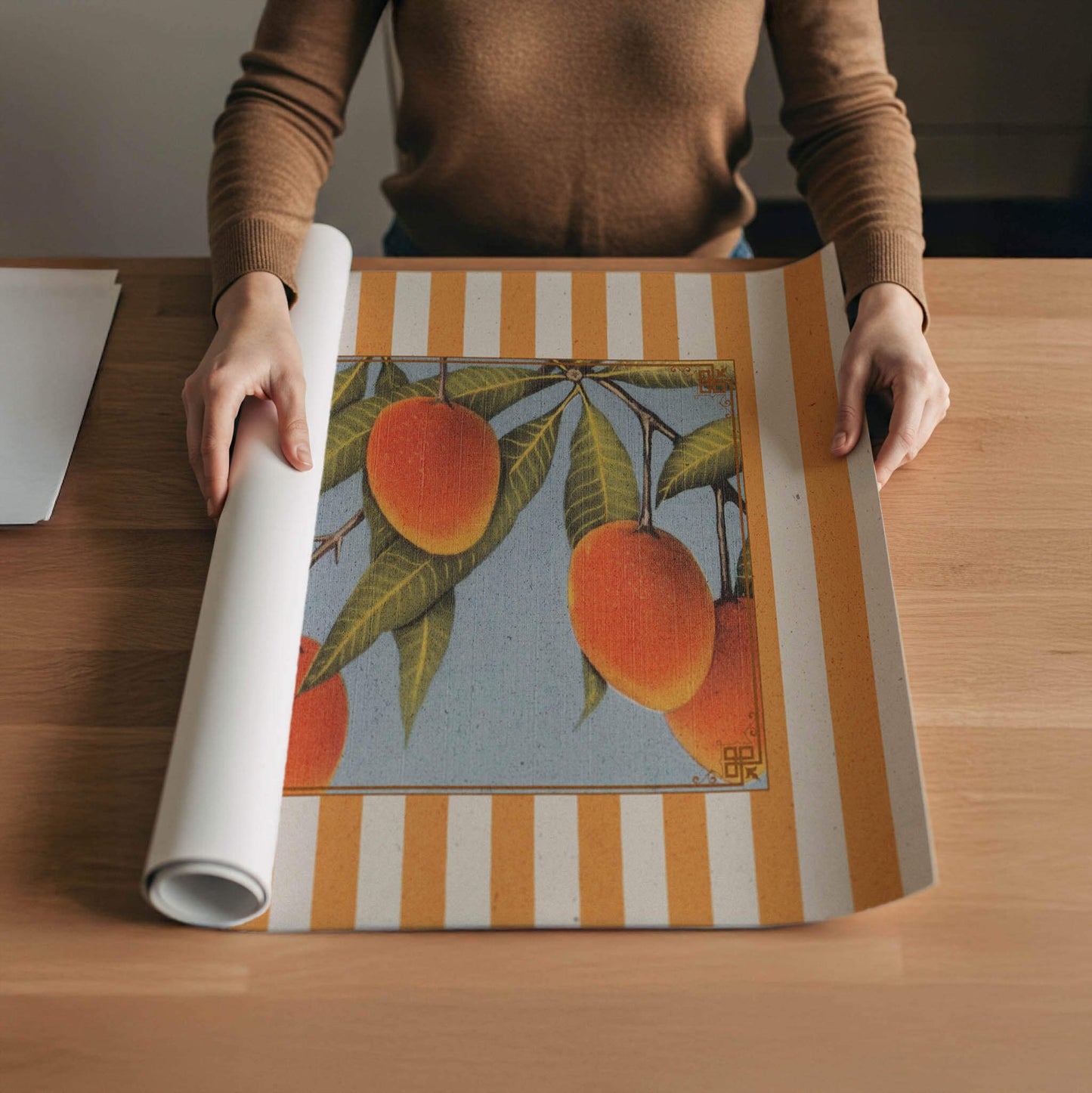 A person is holding a roll of paper with a printed image of oranges on it, which is placed on a wooden table.