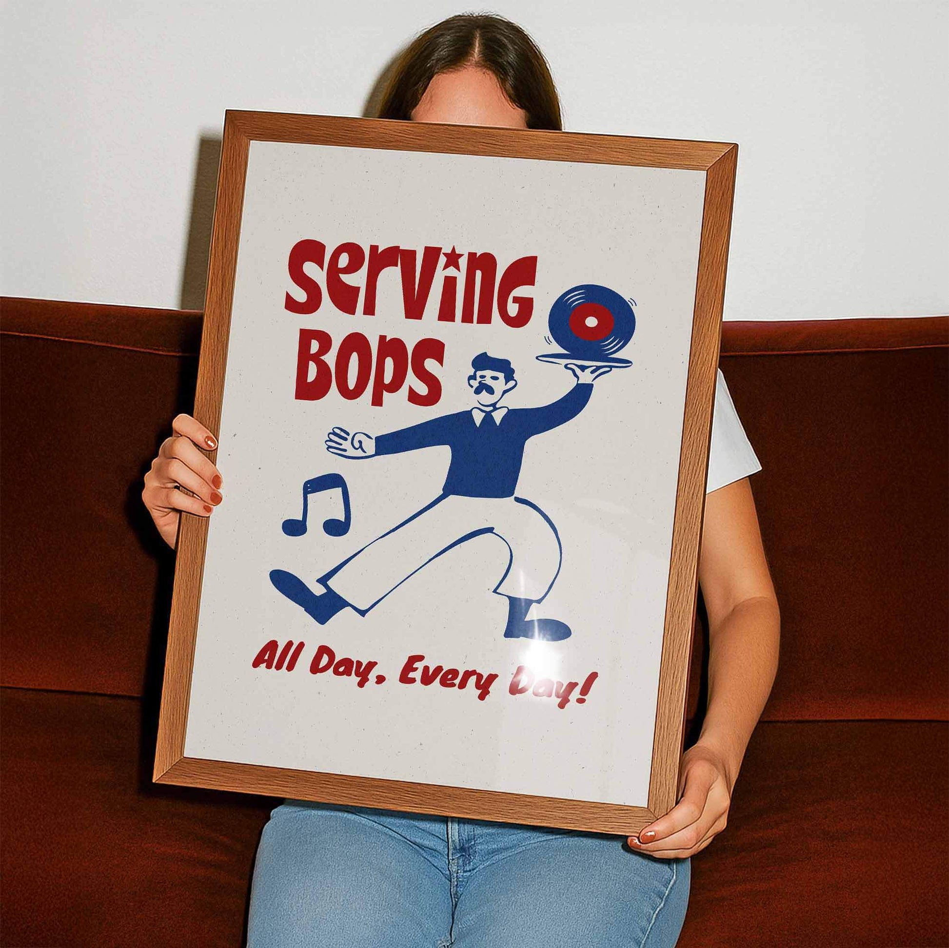 A person is holding up a framed poster with a cartoon illustration of a man serving Bops, accompanied by the text "Serving Bops All Day, Every Day!"