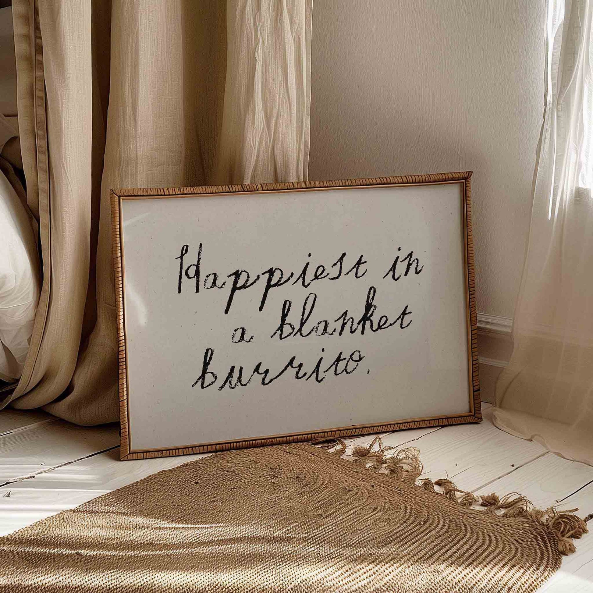 A framed sign with the handwritten text "happiest in a blanket burrito" is placed on a wooden floor next to a beige curtain.