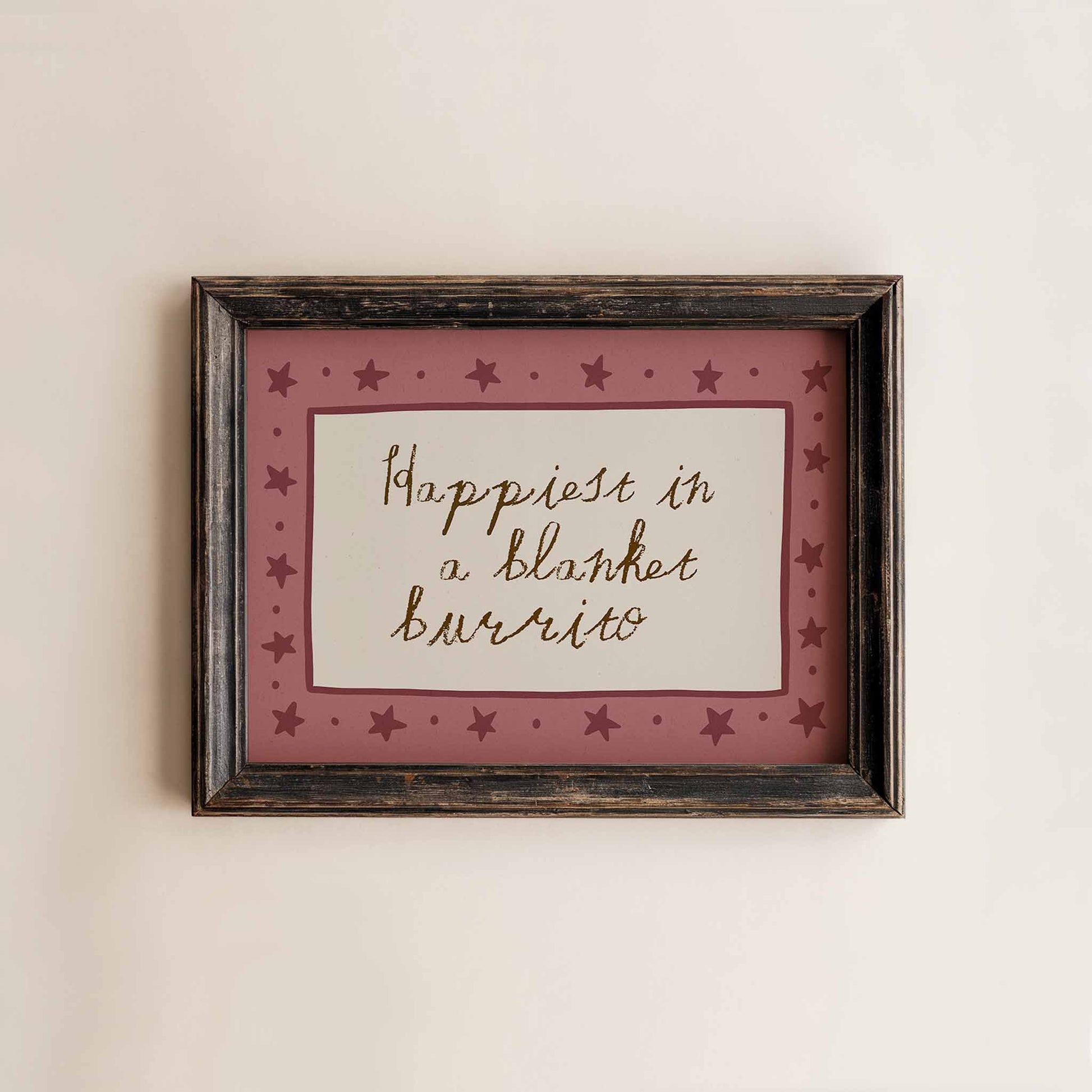 A framed picture with a pink border and stars, containing the handwritten text "Happiest in a blanket burrito".