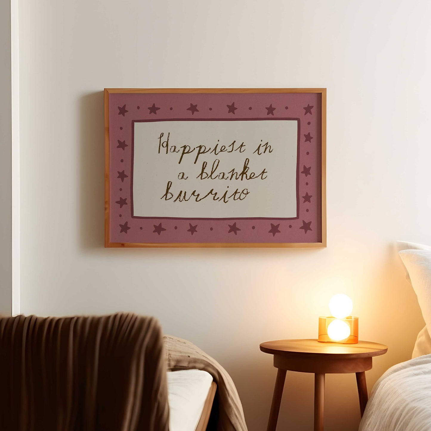 a framed wall art with the text "Happiest in a blanket burrito" in a purple frame with stars. The art is hanging on a white wall above a wooden nightstand with a lamp on it.